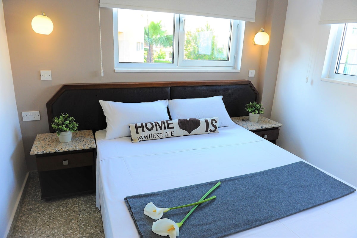 Una cama bien hecha con un cojín que dice “HOME IS WHERE THE HEART IS”, una pequeña maceta sobre la mesita de noche, y una ventana con vistas a la vegetación exterior.