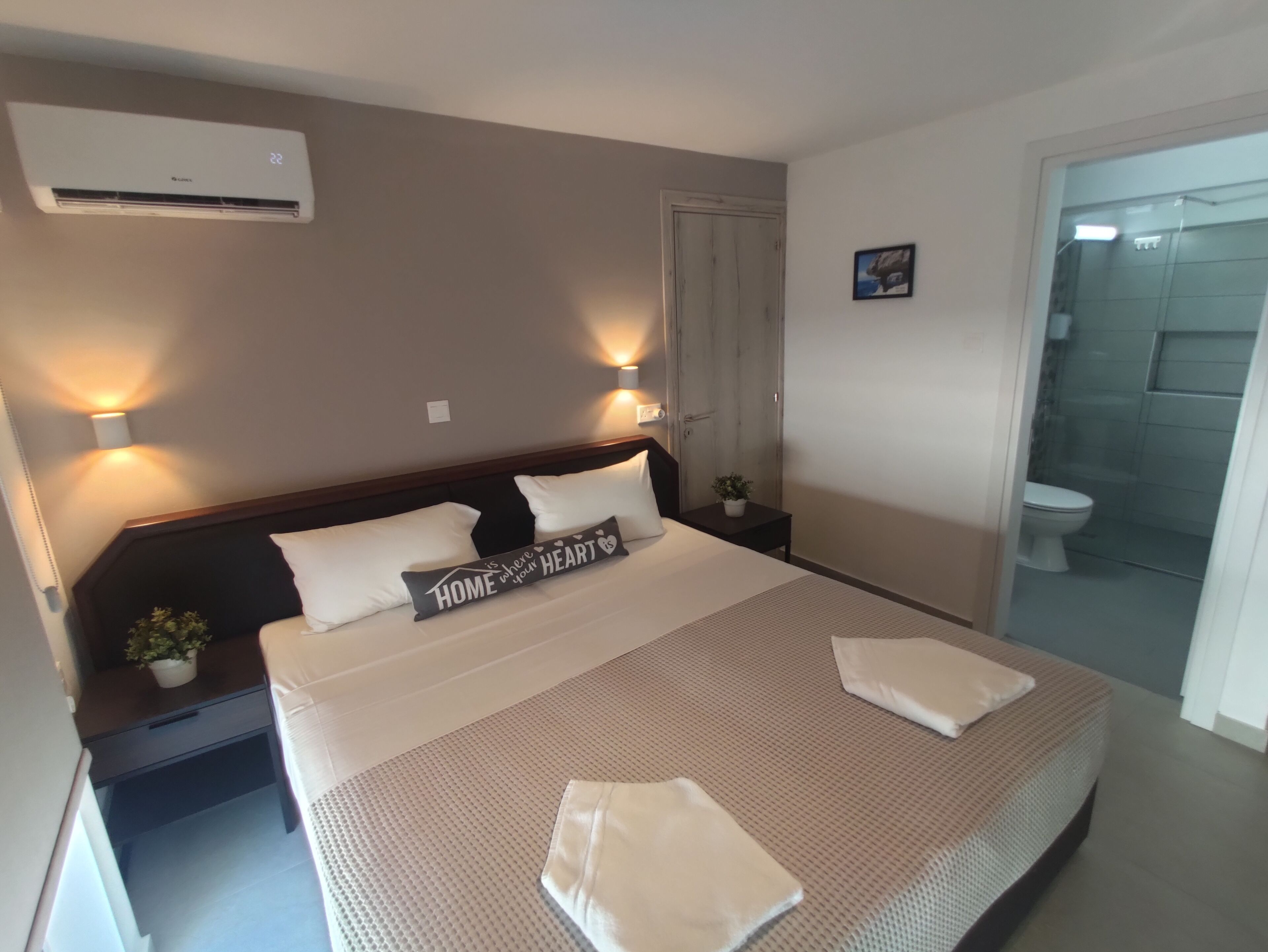 Un dormitorio con cama, mesitas de noche, una planta en maceta, aire acondicionado y un baño visible a través de una puerta abierta.