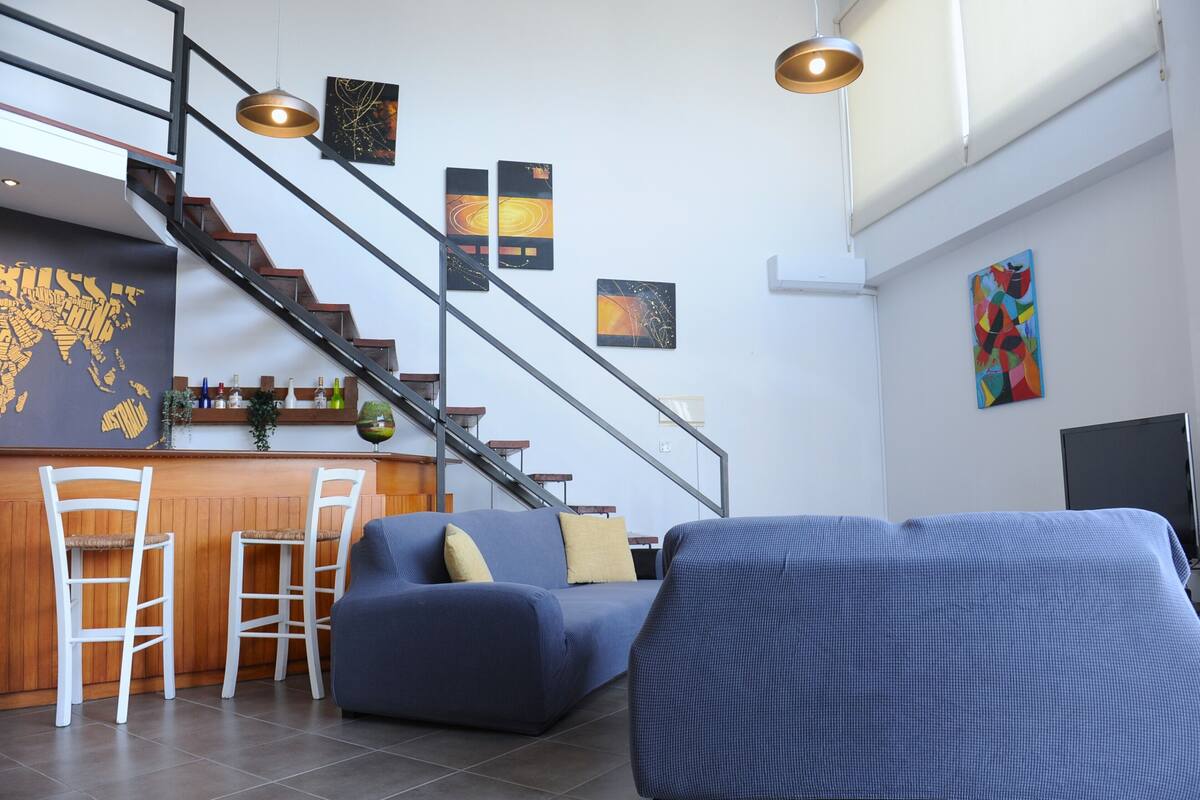 Una sala de estar moderna con un sofá azul, sillas blancas, una zona de bar de madera y una escalera.