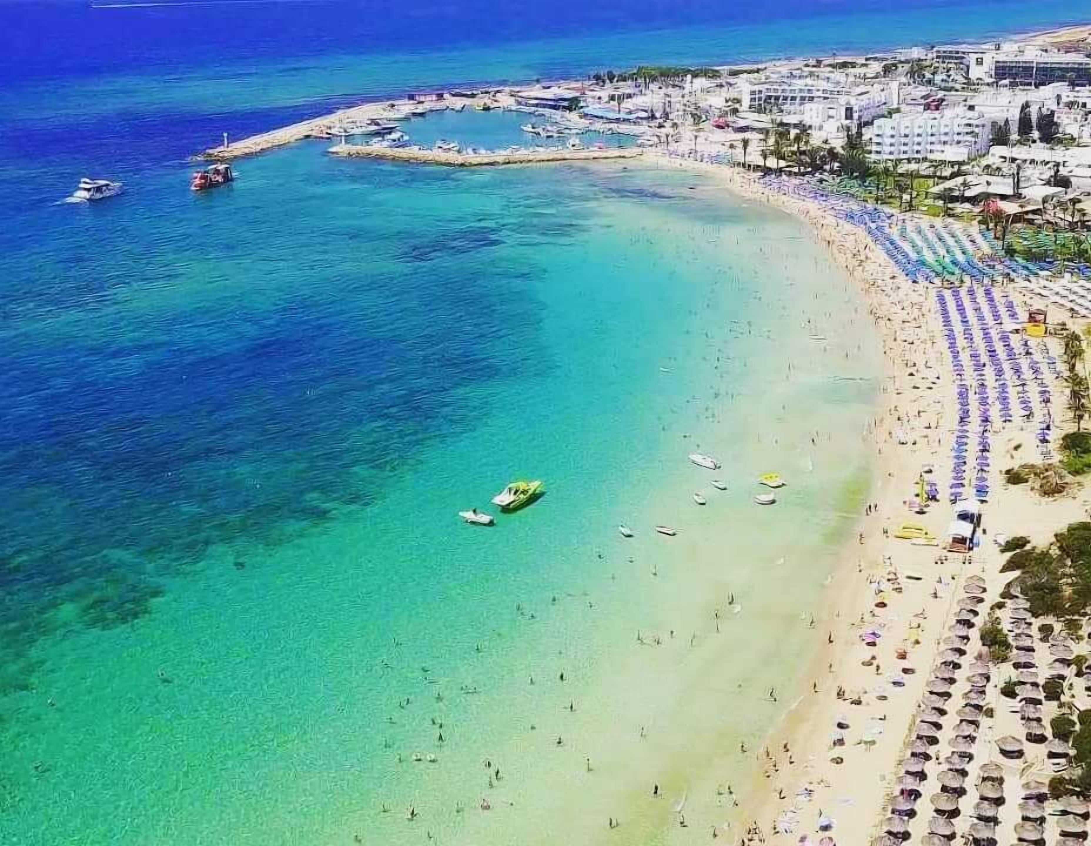A coastal beach with clear turquoise waters, a marina with boats, and a beachfront area with umbrellas and sun loungers.