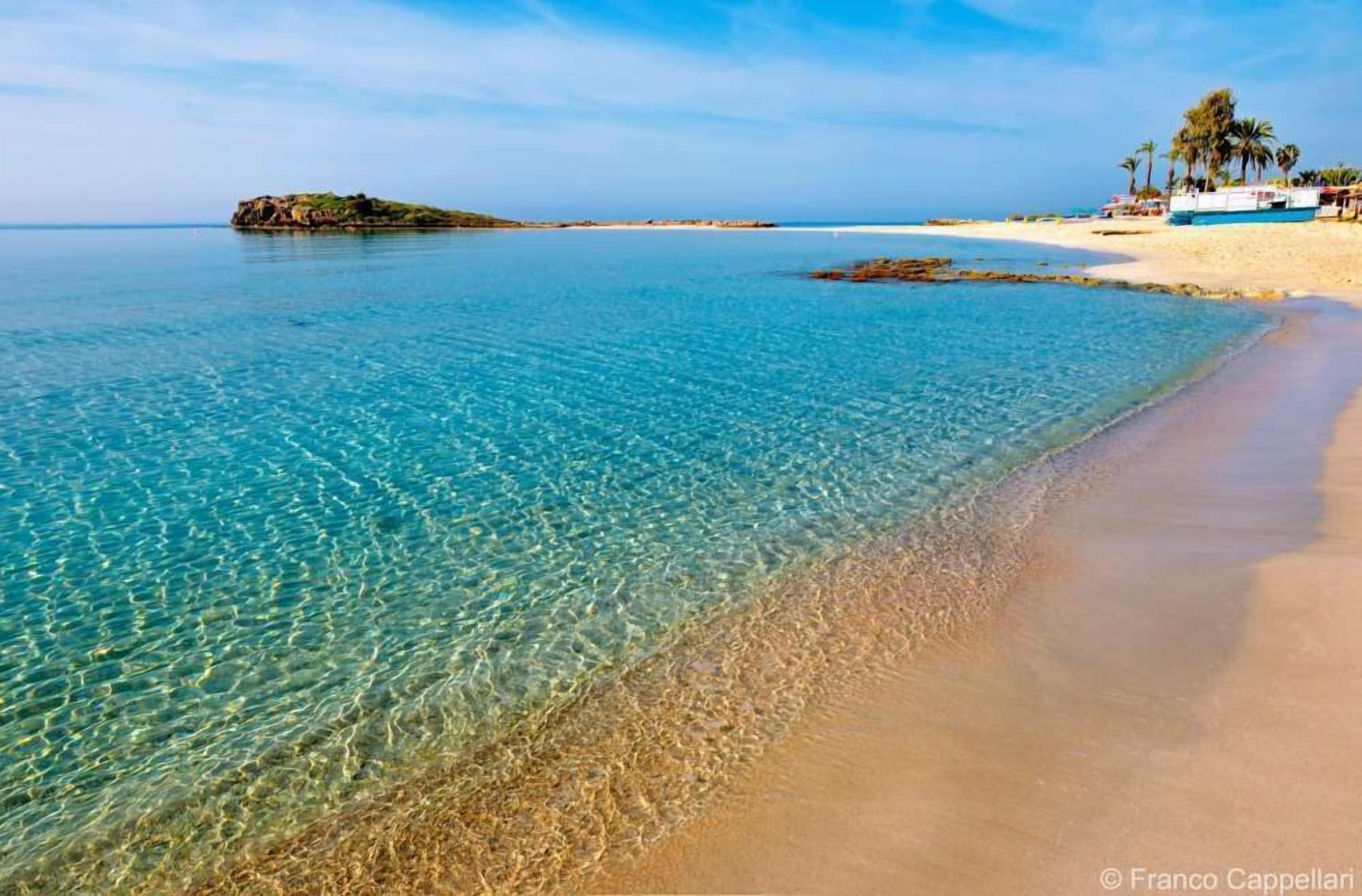 Una playa con agua turquesa cristalina, orilla de arena y una pequeña isla a lo lejos.