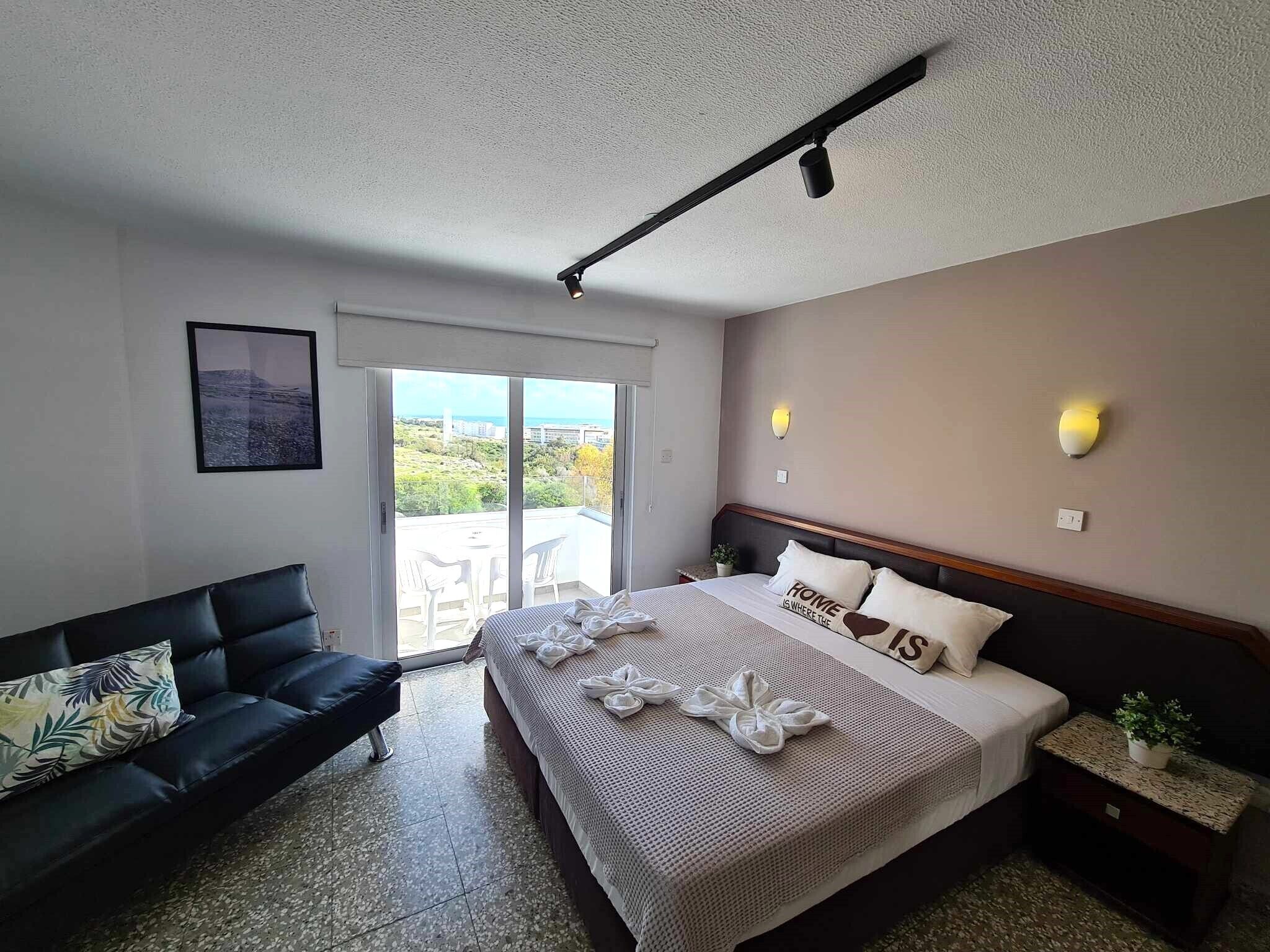 A hotel room with a bed, a sofa, a balcony with chairs, and a ceiling fan.