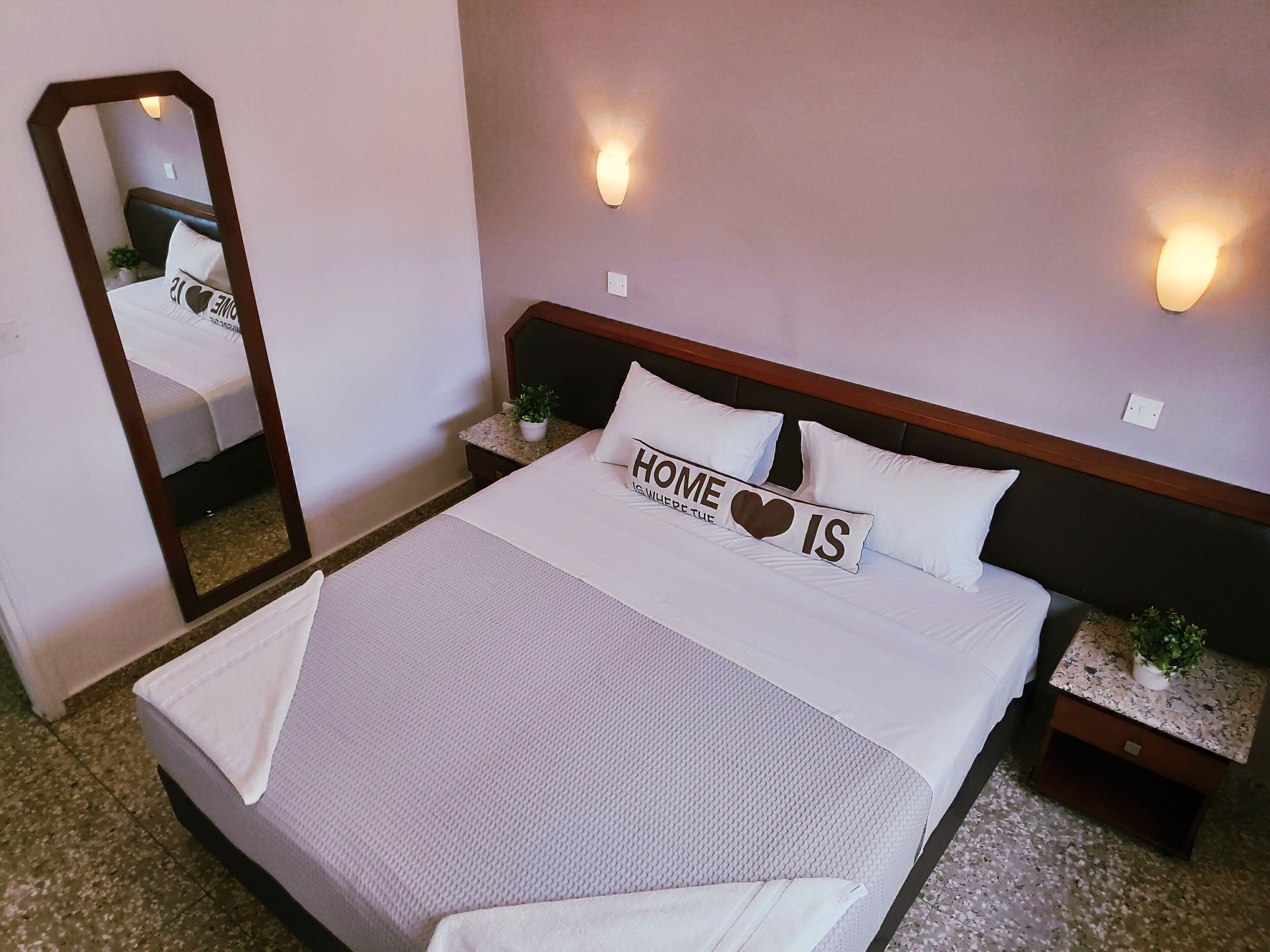 Una cama bien hecha con una almohada que dice 'HOME IS WHERE I AM'. Hay dos luces fijadas en la pared y una planta pequeña en la mesita de noche junto a la cama.