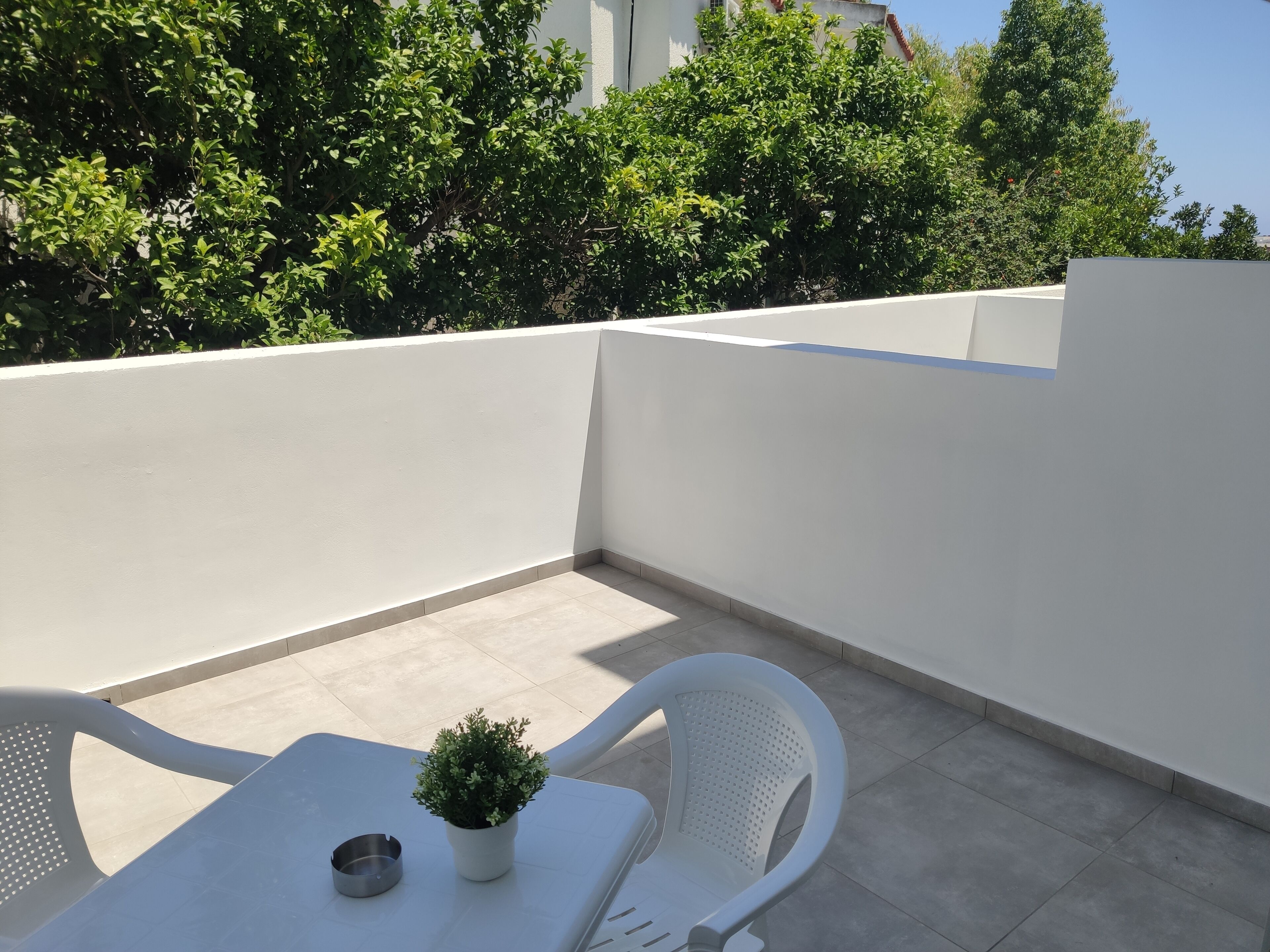 A small outdoor seating area with a white table and two white chairs, a potted plant, and a metal ashtray.