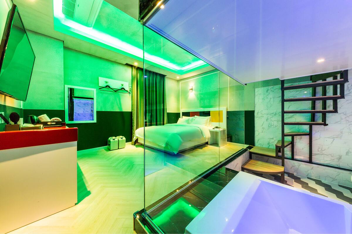 A modern hotel room with a glass partition, a bed, a bedside table, a nightstand, a desk, a chair, a desk lamp, a telephone, a television, and a bathtub.