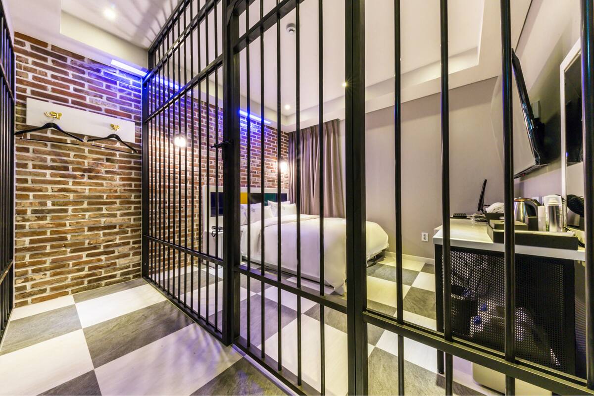 A hotel room with a checkered floor, a bed, a desk with a computer, a TV, and a brick wall with a hanging garment rack.