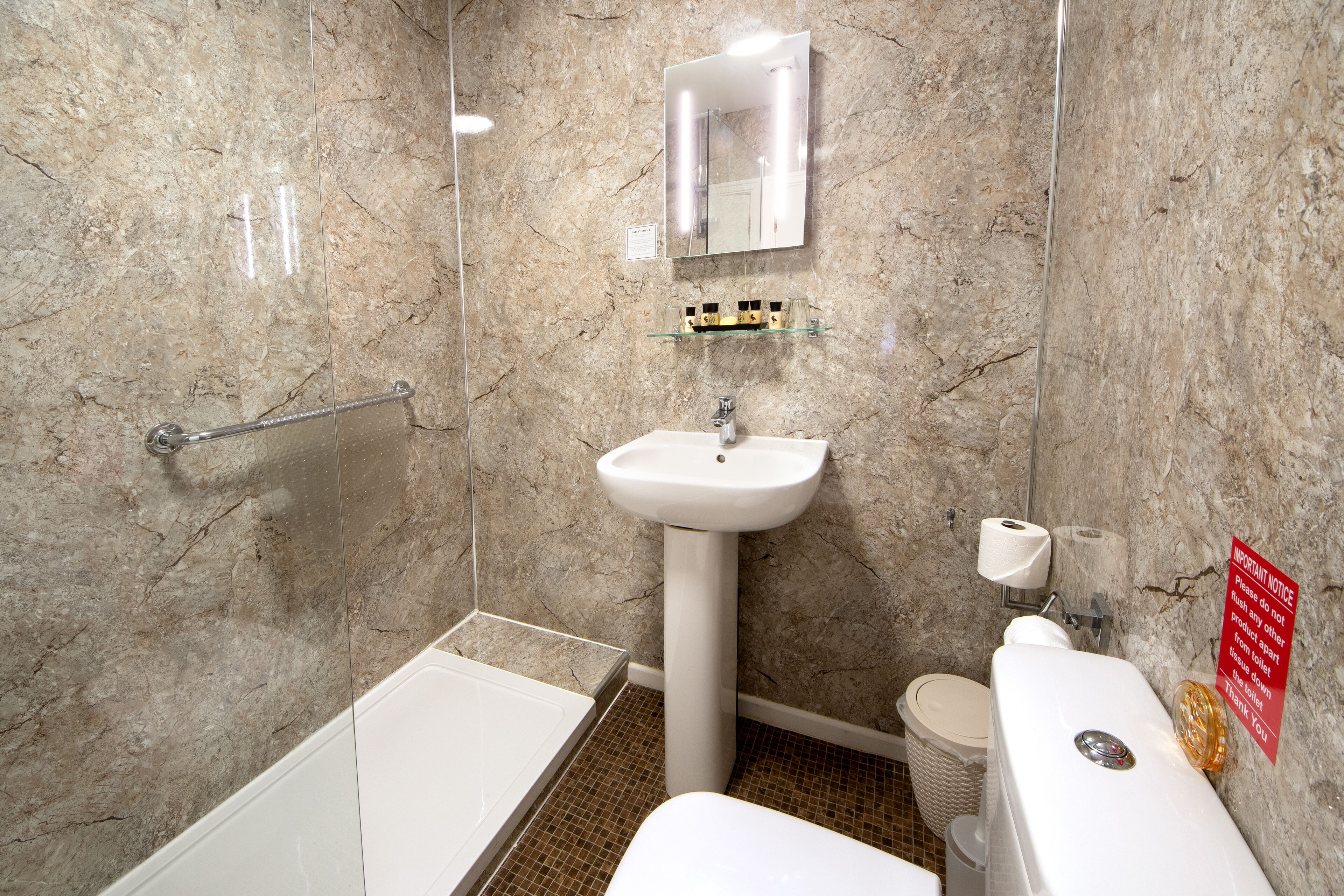 Un baño con pared de mármol, un lavabo blanco, estante de vidrio, baranda y un inodoro.