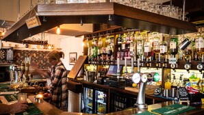 A bar area with a bartender, shelves of liquor bottles, and a cash register. - Maypole Inn (Skipton)