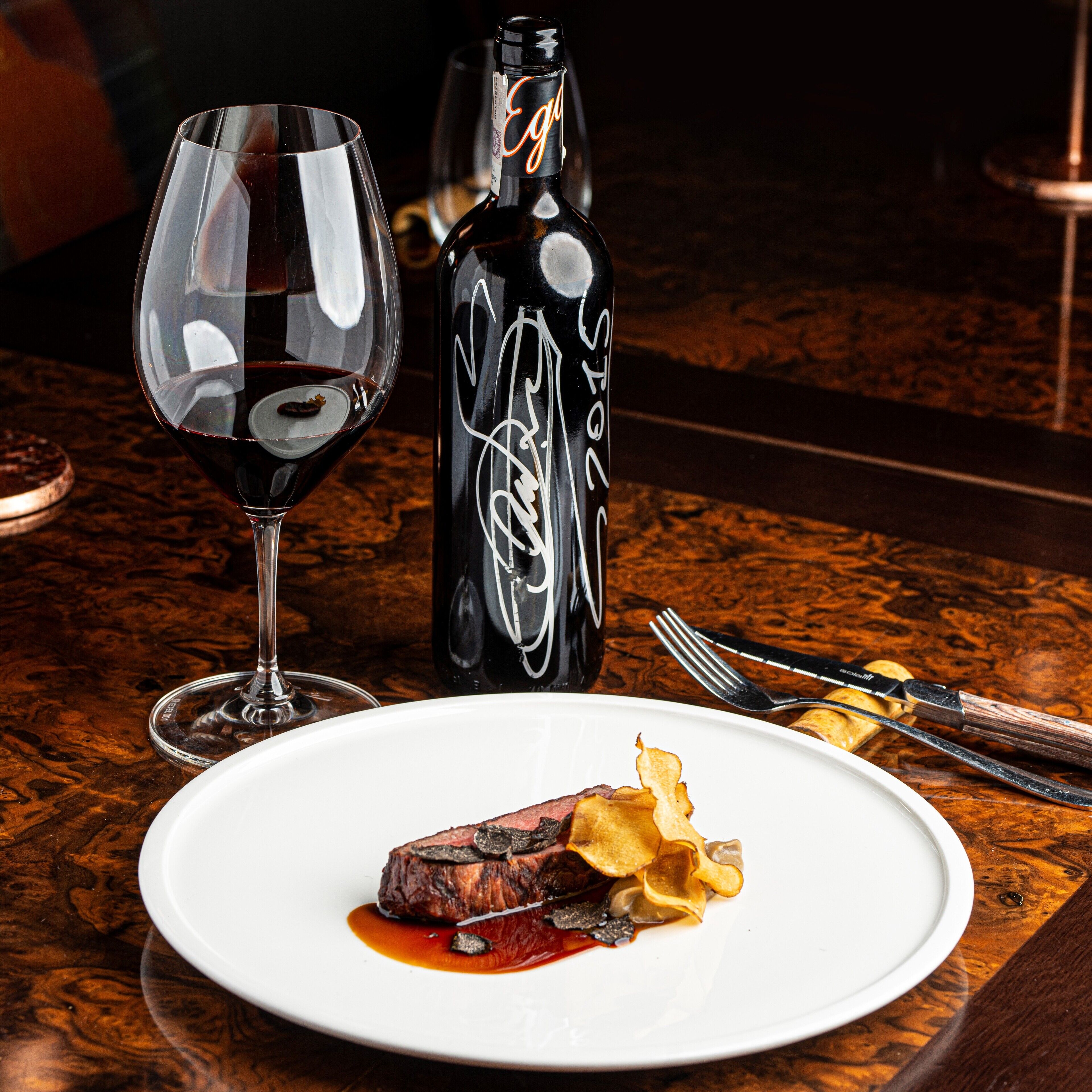 Un plato con carne, papas fritas, una copa de vino y una botella de vino sobre una mesa de madera.
