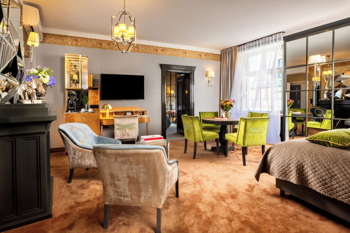 A hotel room with a large bed, two armchairs, a dining table, a TV, and a mirror.