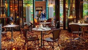 Un restaurant avec des tables rondes et des chaises en osier, un sol carrelé et une vue sur la cuisine à travers une porte vitrée.