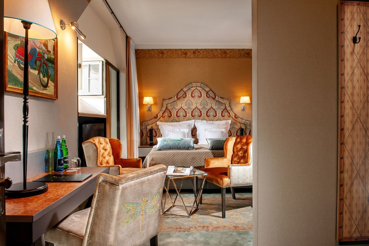 A hotel room with a bed, two chairs, a desk, a lamp, and a painting.