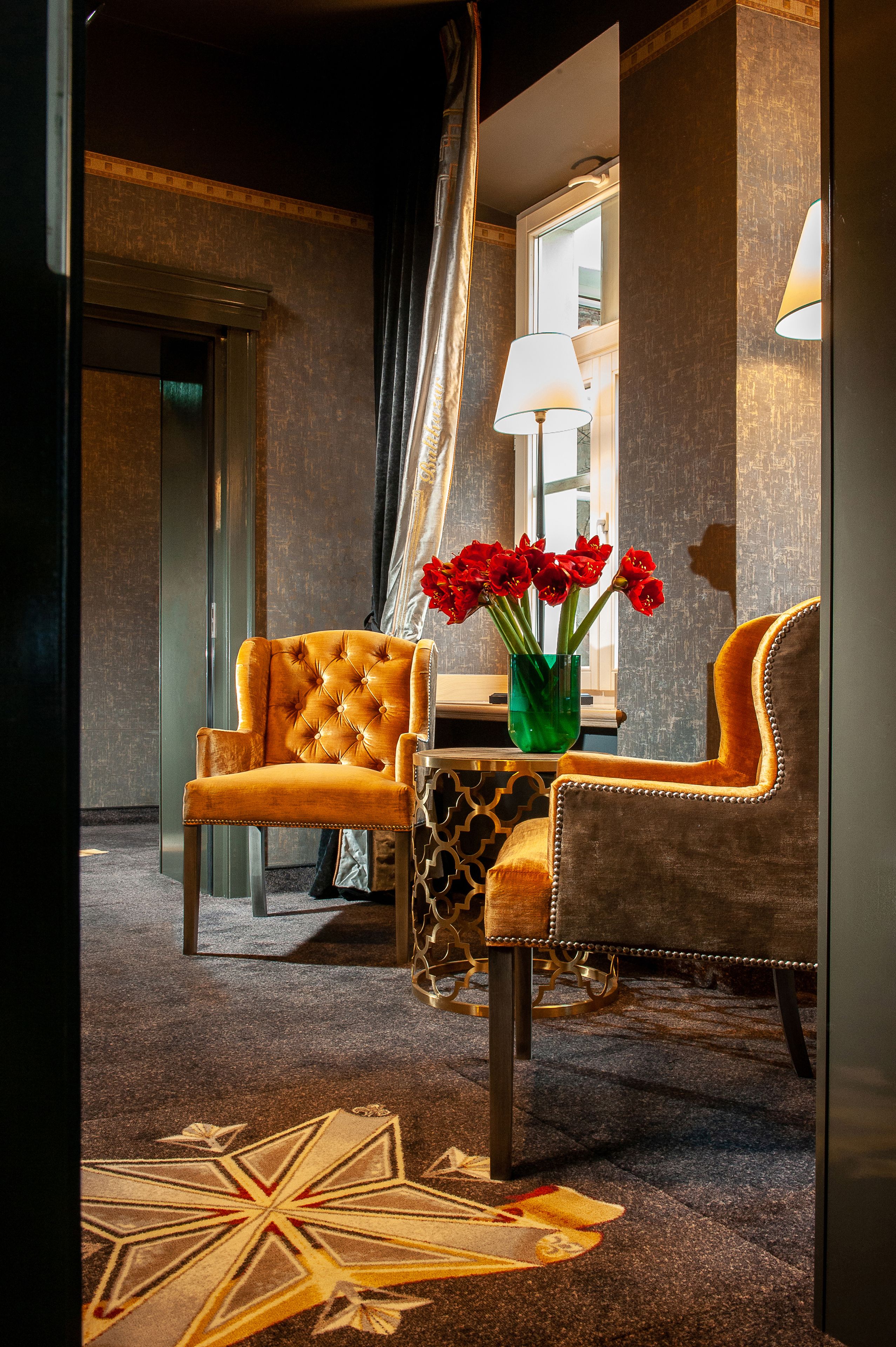 A hotel room with a tufted armchair, a side table with a vase of red flowers, and a patterned rug.