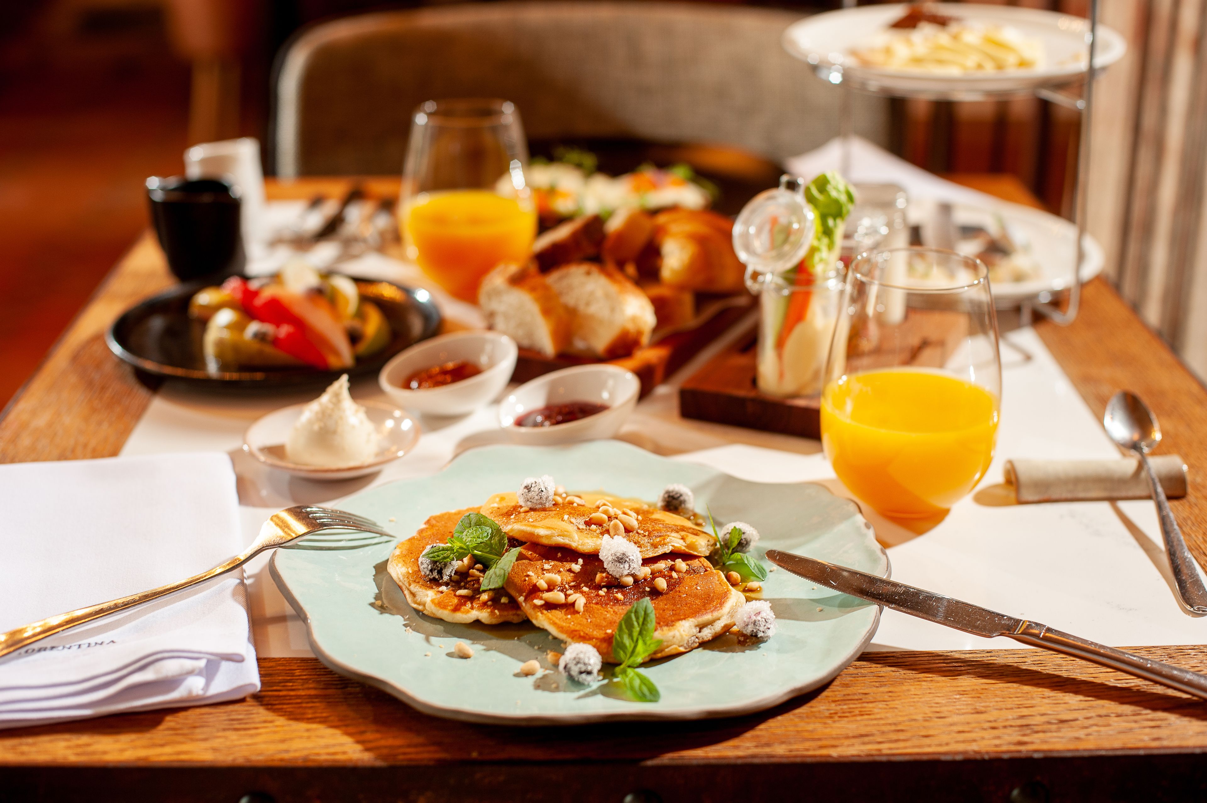 Une table en bois dressée pour le petit-déjeuner, avec des crêpes, du jus d’orange et divers accompagnements.