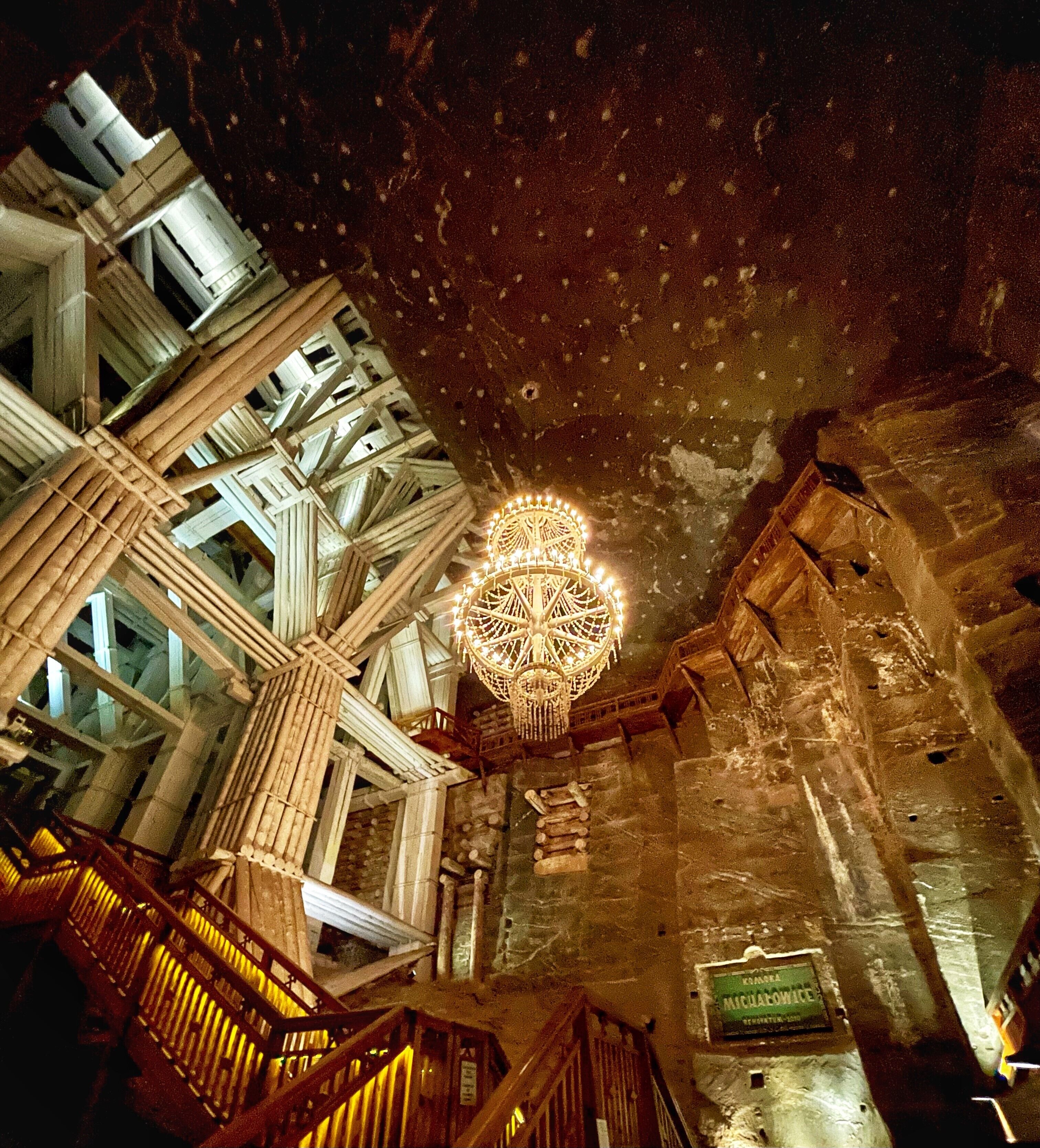 An interior view of a historic building with a large, ornate chandelier hanging from the ceiling, a stone wall with a plaque, and a wooden staircase.