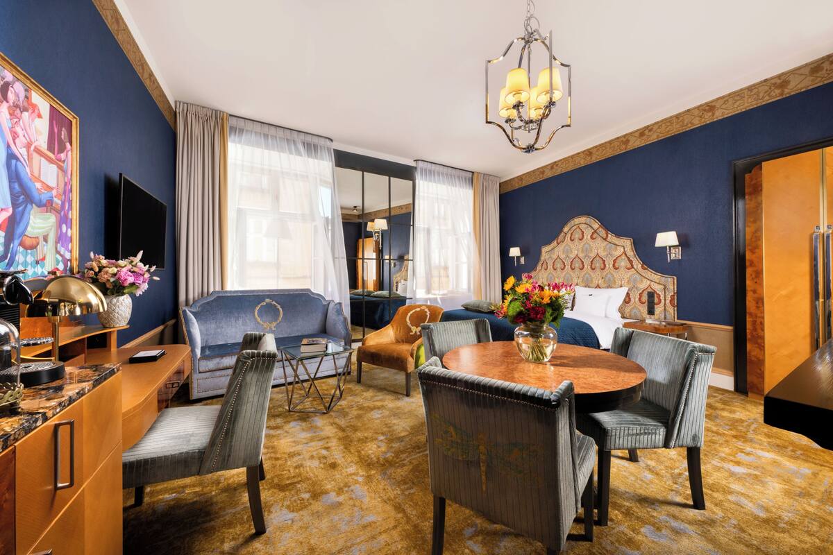 A hotel room with a large bed, a sitting area with a sofa and chairs, a dining table with chairs, a TV, and a chandelier.
