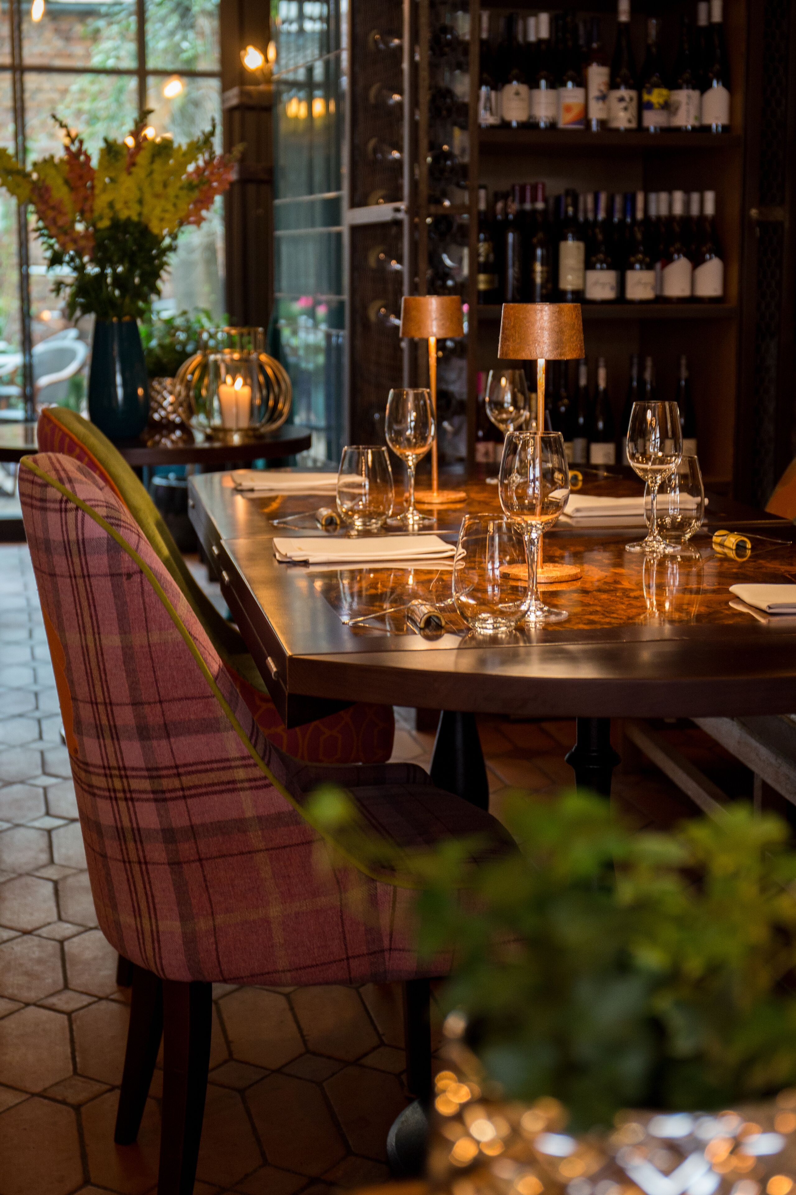 Een restaurant met een lange tafel gedekt voor diner, een wijnrek op de achtergrond en een raam met uitzicht op buiten.