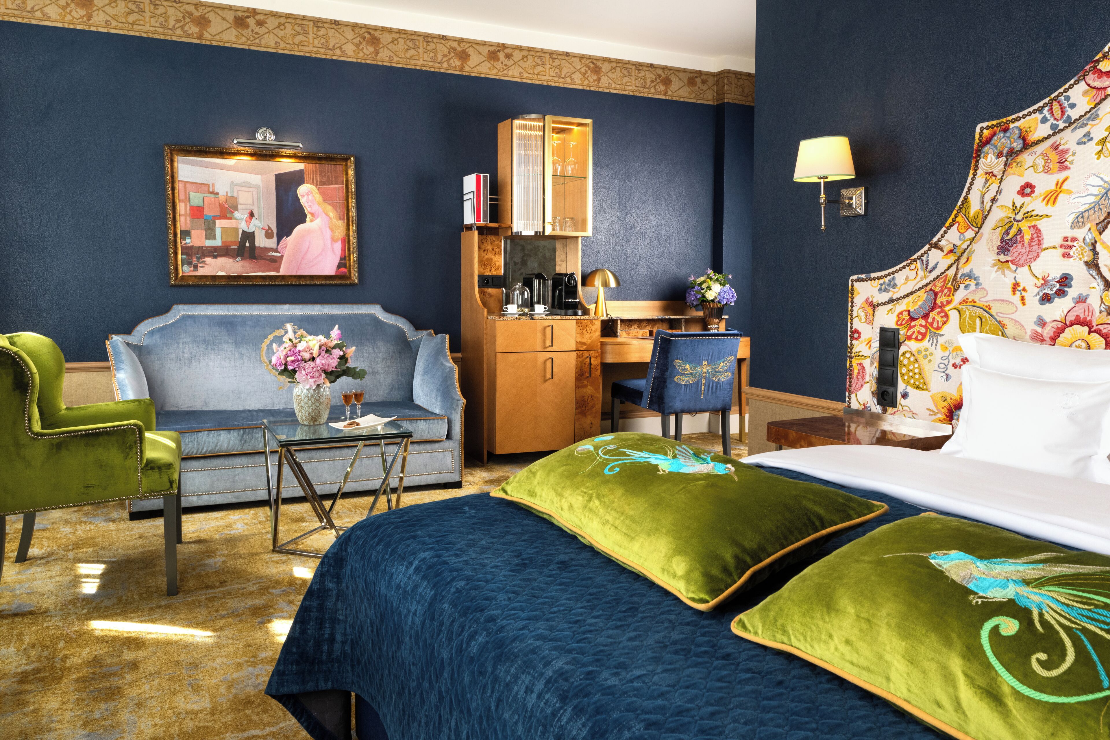 A hotel room with a bed, two green armchairs, a sofa, a desk with a lamp, a coffee maker, and a painting on the wall.