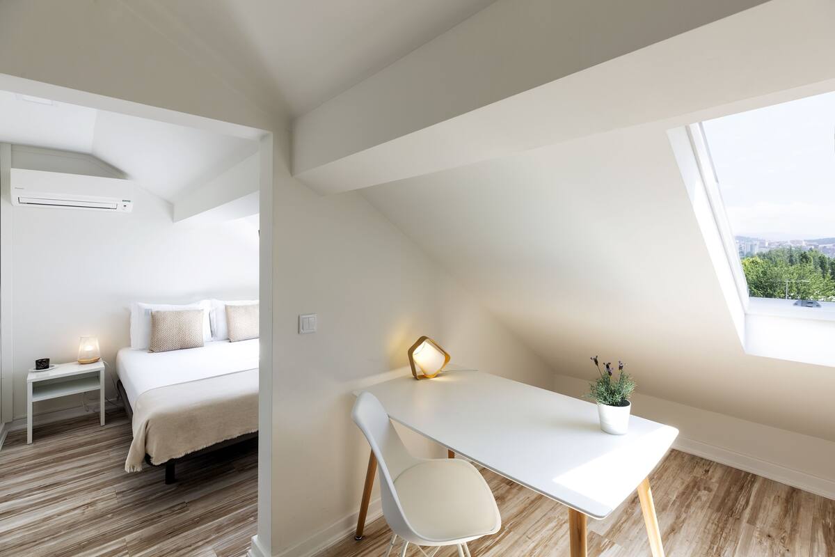 A modern bedroom with a bed, bedside table, and a desk with a chair. There is a window providing a view of greenery and a cityscape.