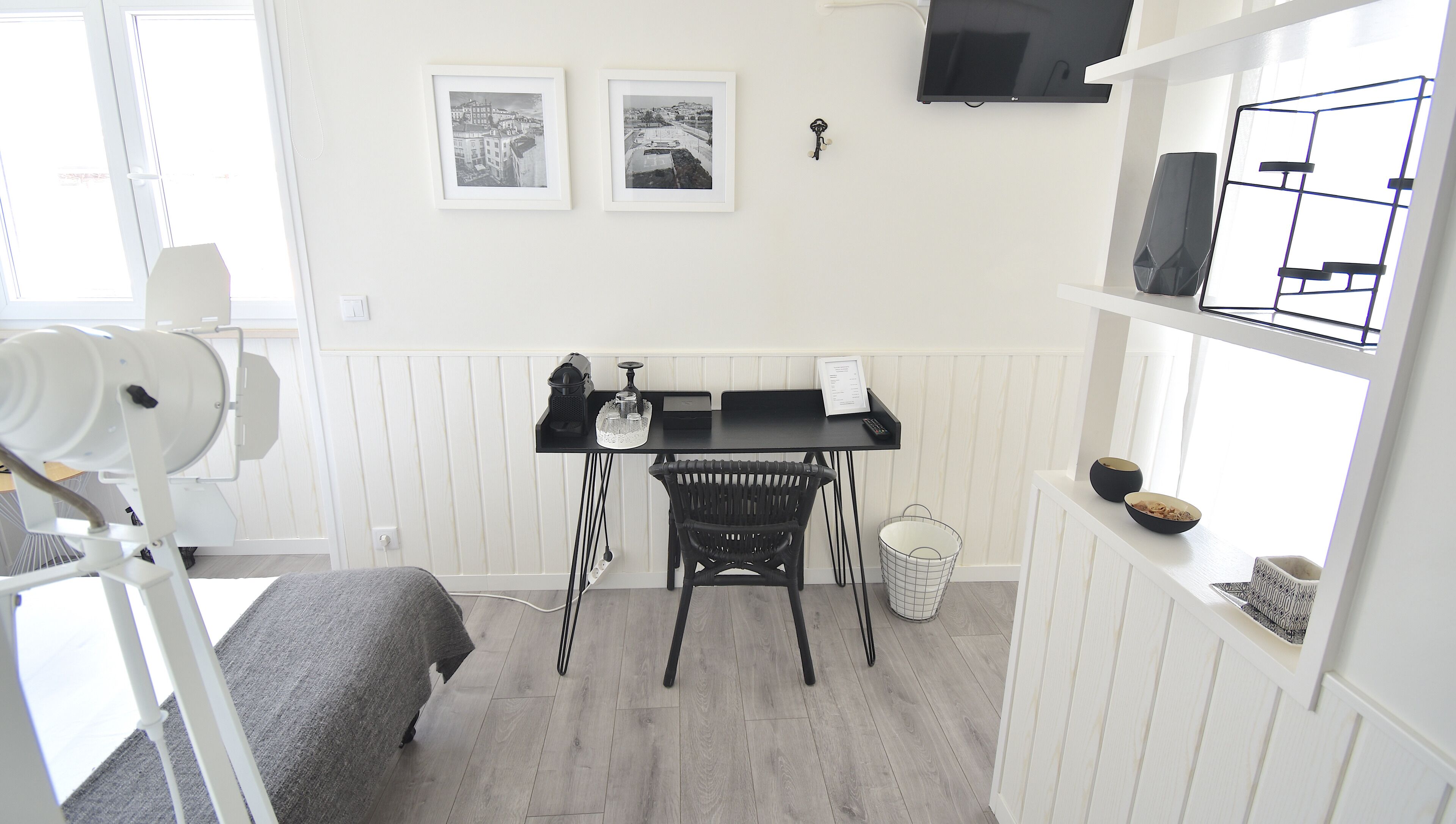 A small, well-lit room with a desk, chair, and a television mounted on the wall. There are two framed pictures on the wall above the desk.