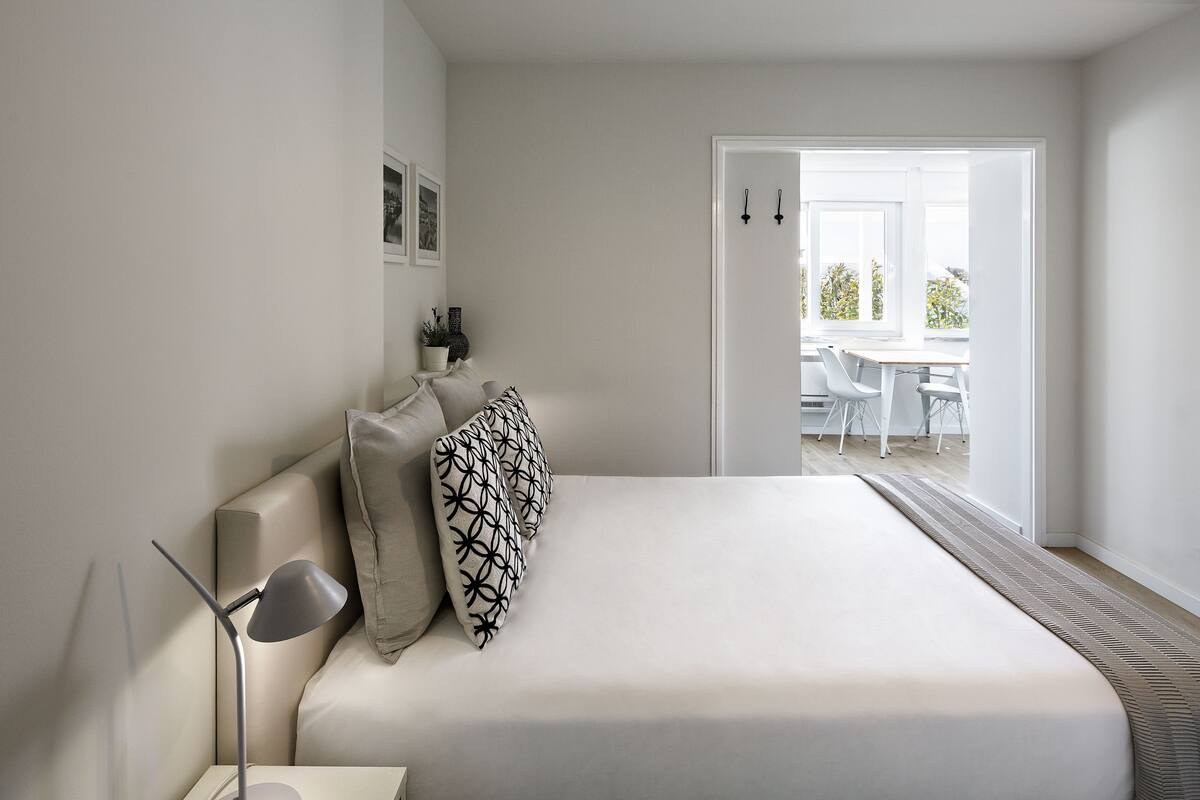 A minimalist bedroom with a large bed, a bedside table, and a nightstand. There is a door leading to a balcony with outdoor furniture.