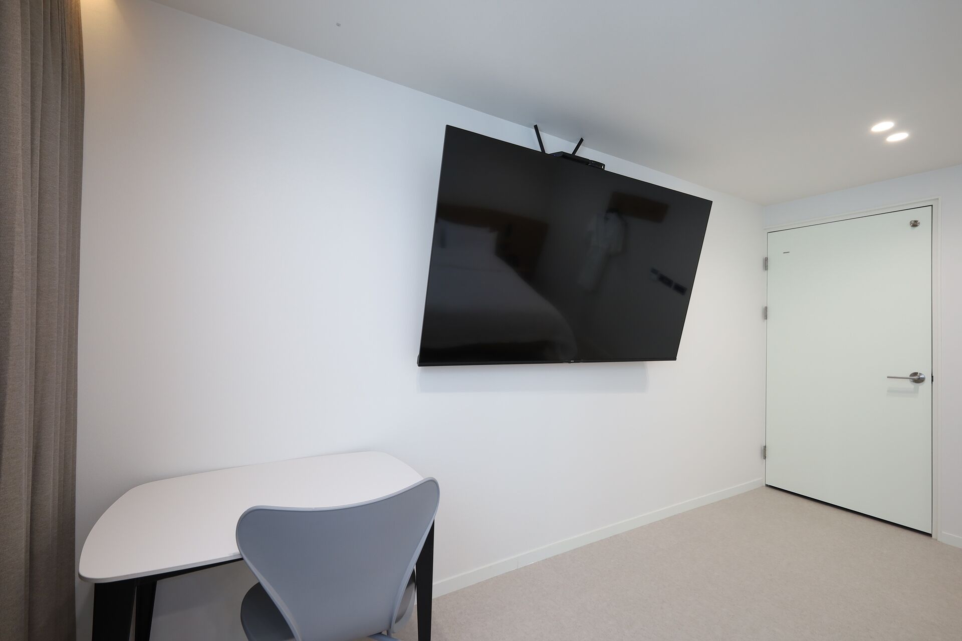 Una habitación moderna con una gran televisión de pantalla plana montada en la pared, una puerta blanca y una pequeña mesa blanca con una silla gris.