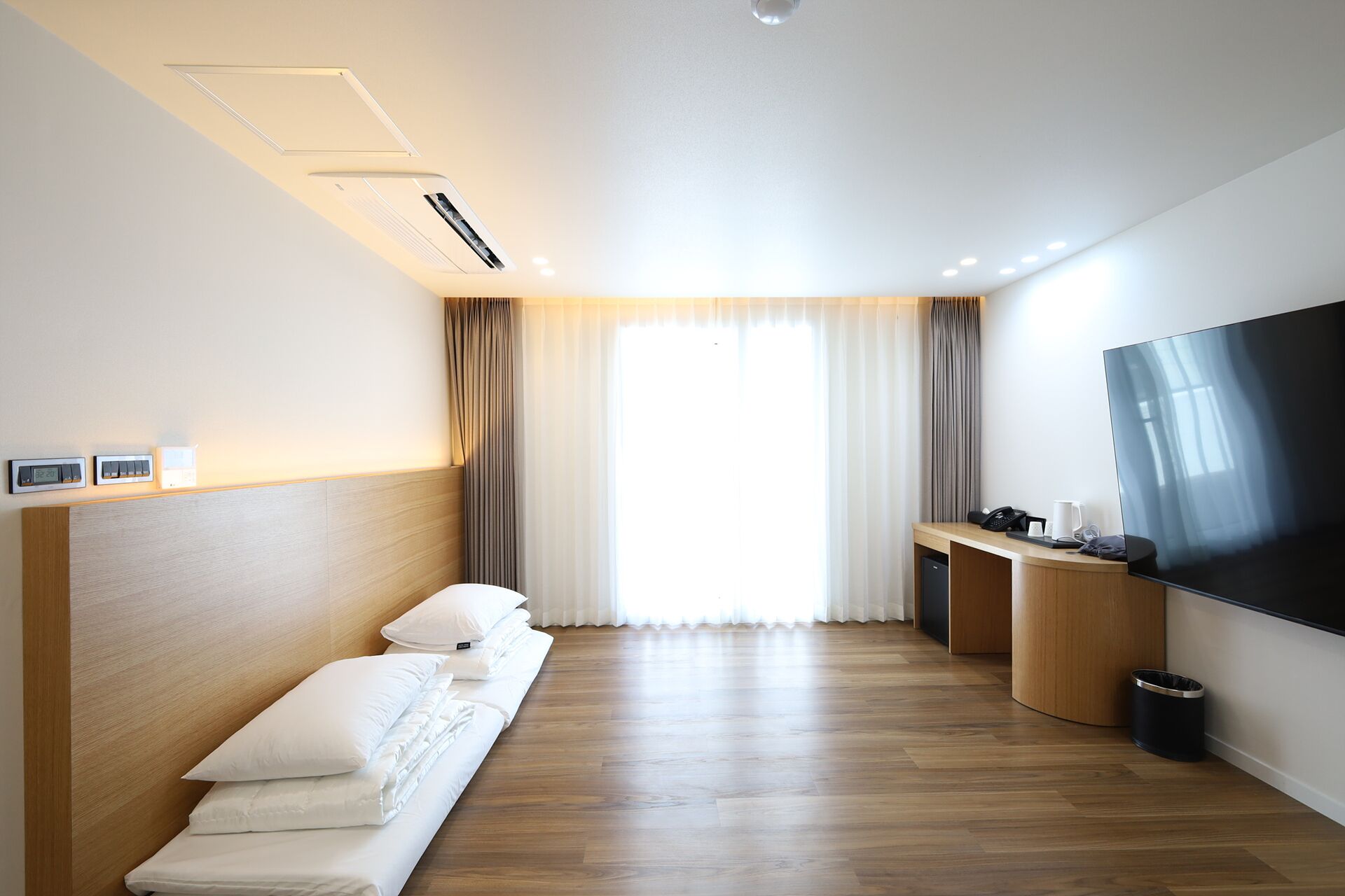 Habitación de hotel con piso de madera, una cama con sábanas blancas, un televisor en la pared, un escritorio con un teléfono y una taza, y una ventana con cortinas.