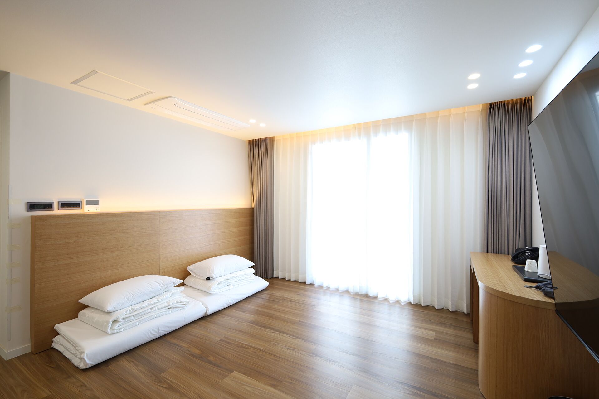 Quarto de hotel com cabeceira de madeira, cama com roupa de cama branca, escrivaninha com telefone e uma janela grande com cortinas.