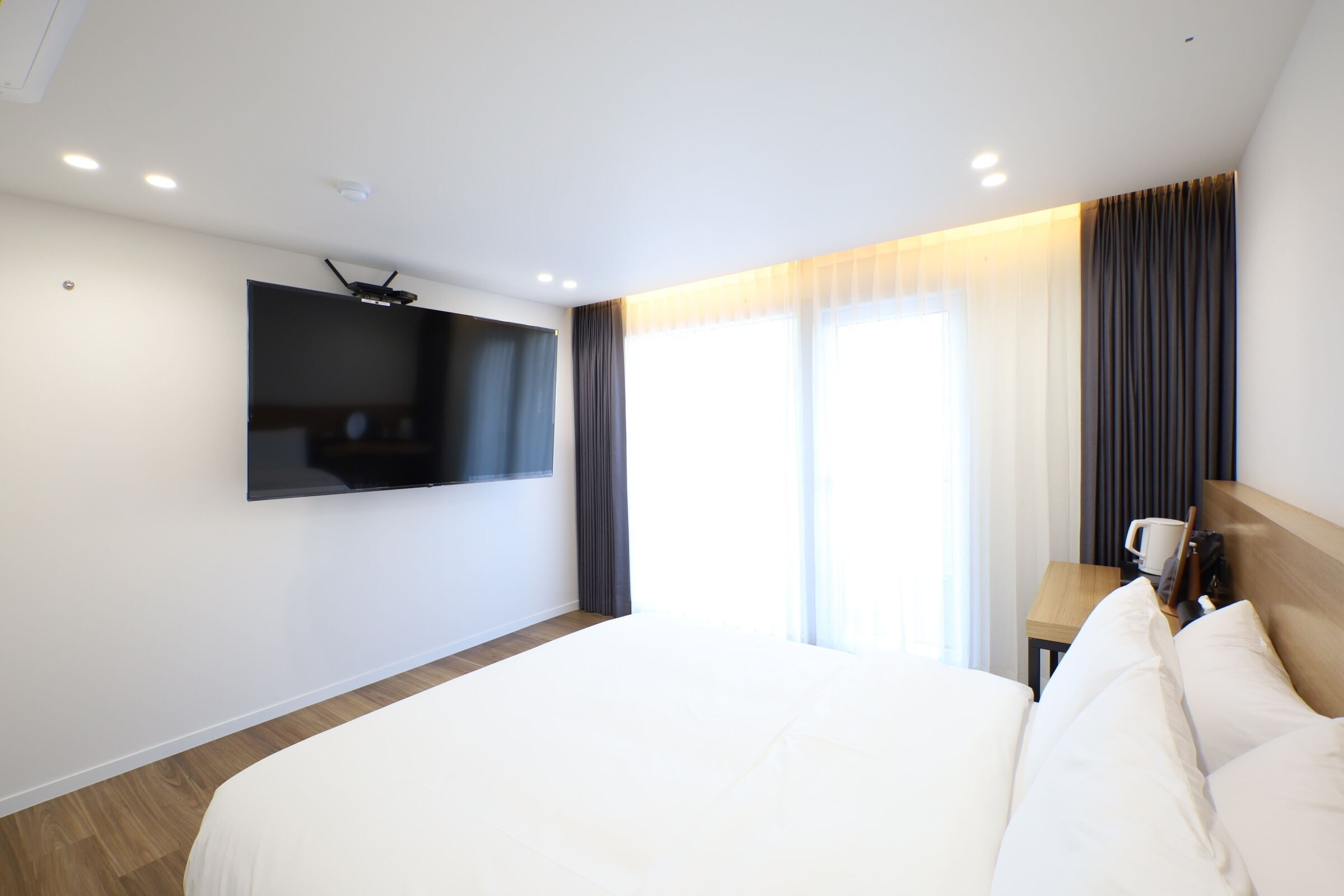 A hotel room with a large bed, a flat-screen TV mounted on the wall, a small table with a cup and a book, and a window with curtains.