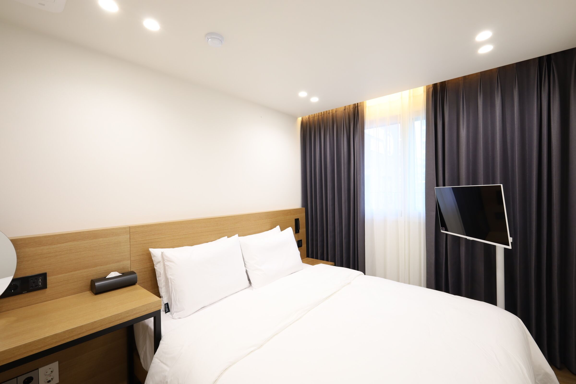 A hotel room with a neatly made bed, a bedside table with a lamp, and a large window with curtains.