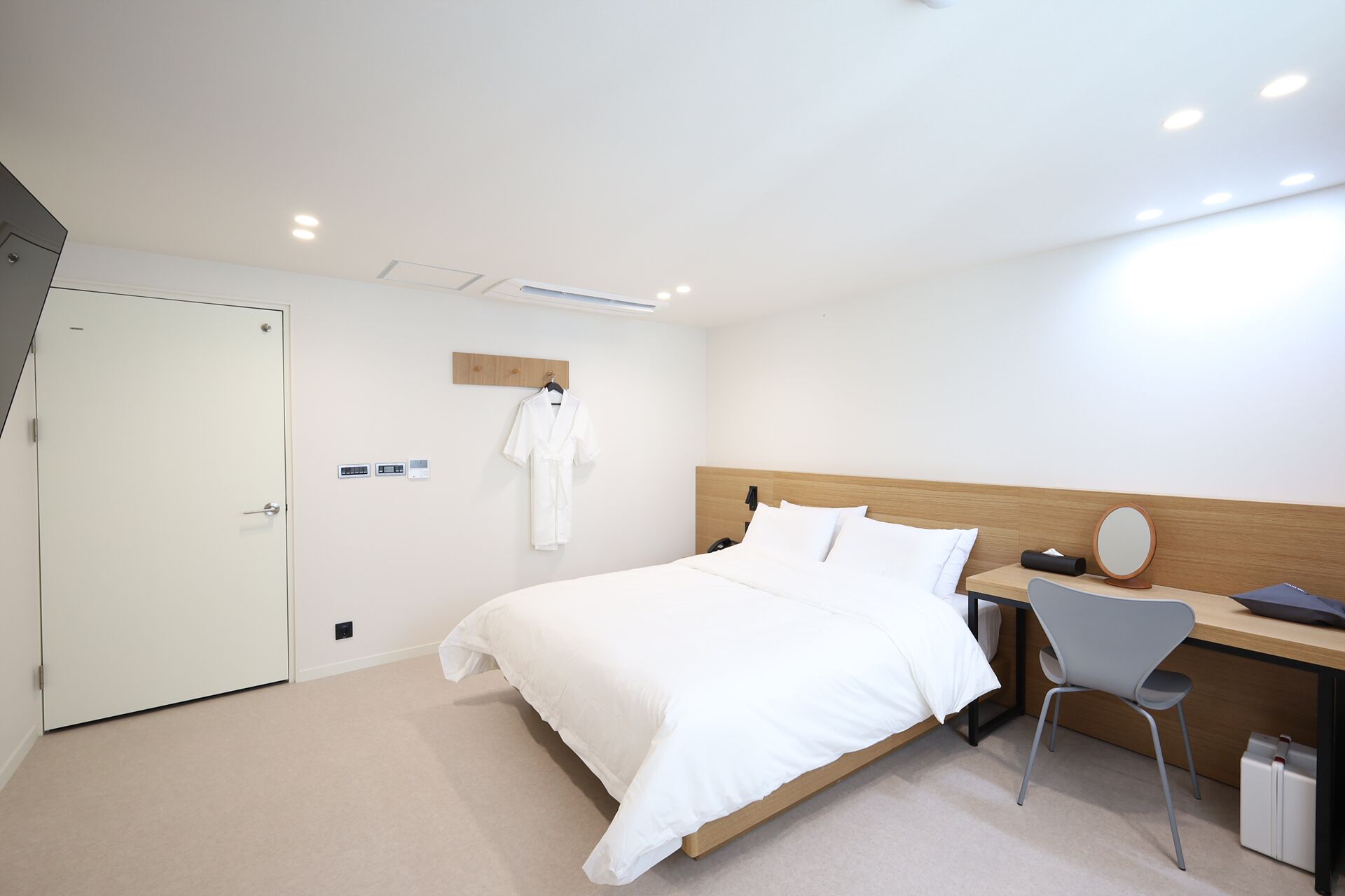 Quarto de hotel moderno com cama, escrivaninha com cadeira, roupão pendurado na parede e cabeceira de madeira.