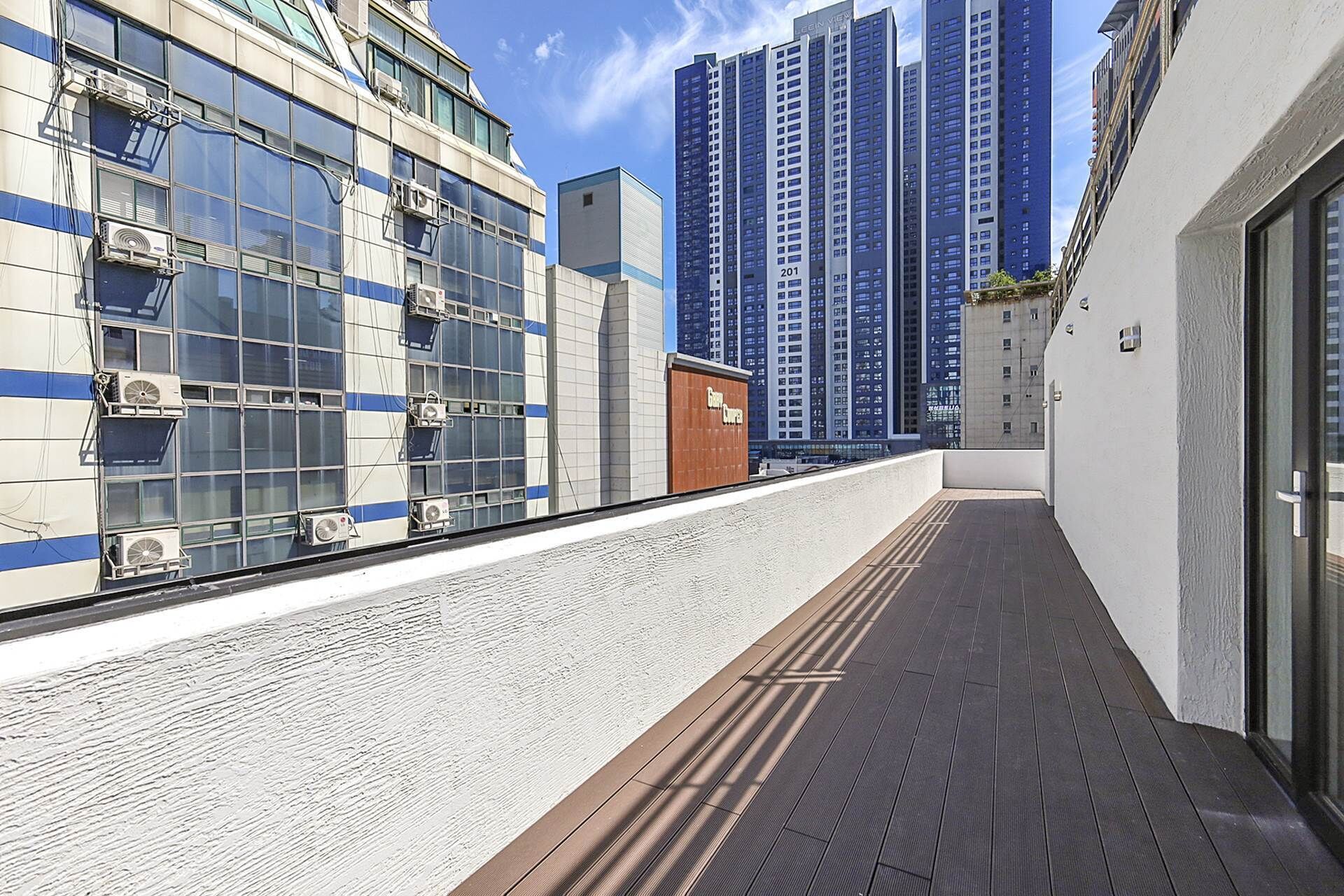 Un edificio moderno con amplios ventanales, unidades de aire acondicionado, una terraza de madera y rascacielos al fondo.