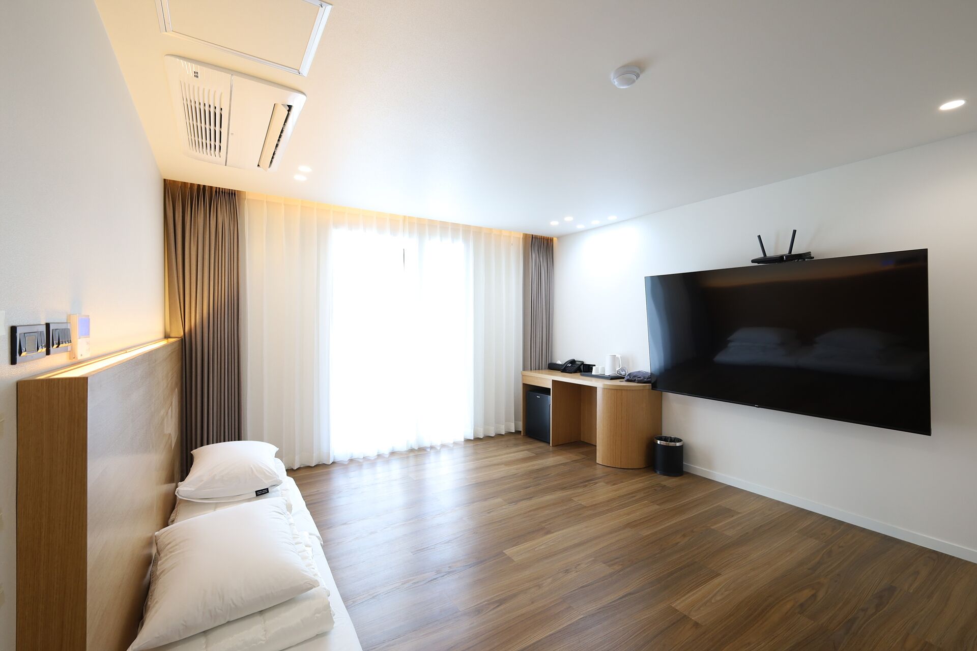 A modern hotel room with a wooden floor, a large flat-screen TV mounted on the wall, a desk with a chair, a sofa with cushions, and a window with curtains.