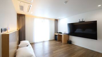 A modern hotel room with a wooden floor, a large flat-screen TV mounted on the wall, a desk with a chair, a sofa with cushions, and a window with curtains.