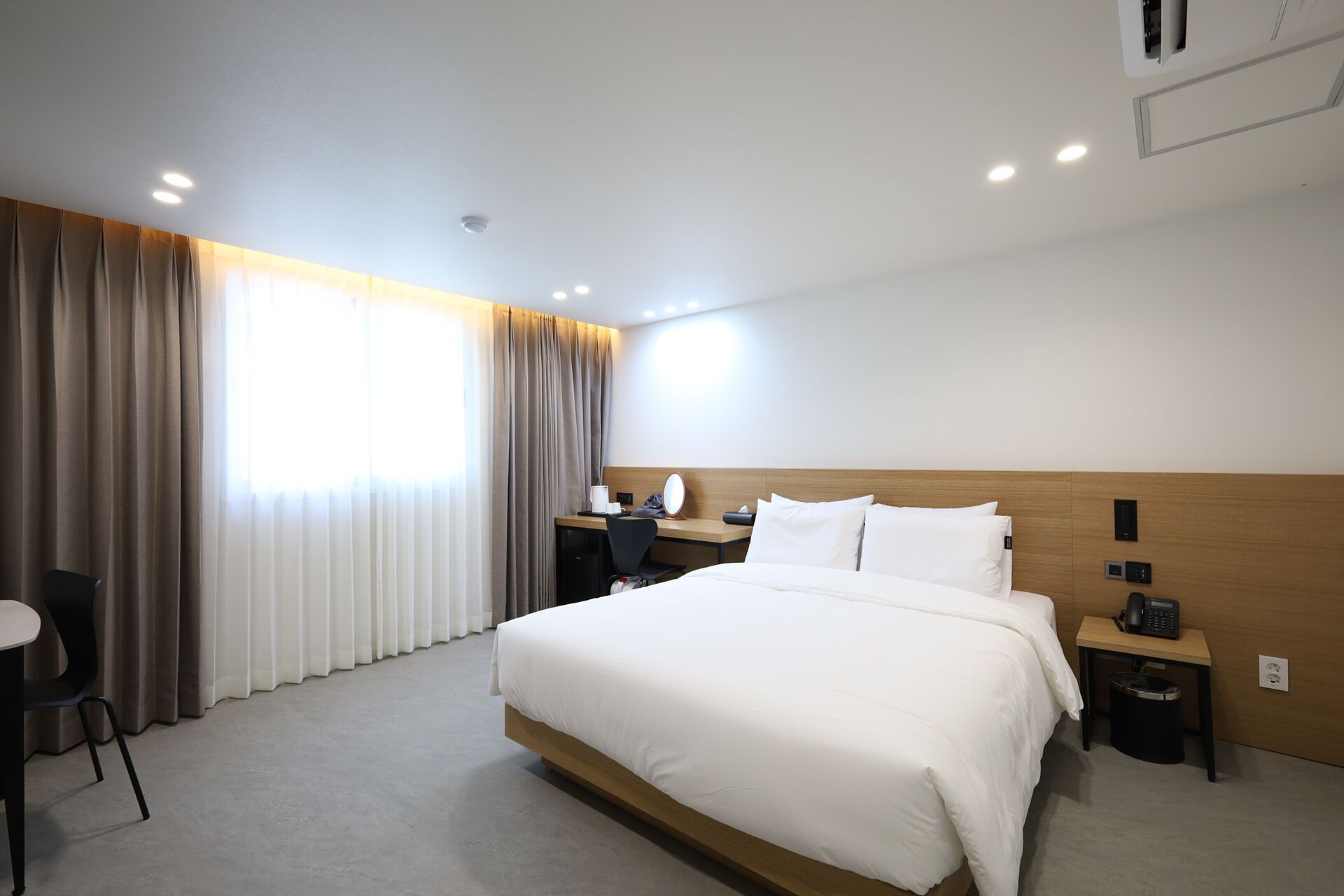 Ein Hotelzimmer mit einem großen Bett, einem Schreibtisch mit Stuhl, einem Nachttisch mit Telefon und einem Fenster mit Vorhängen.