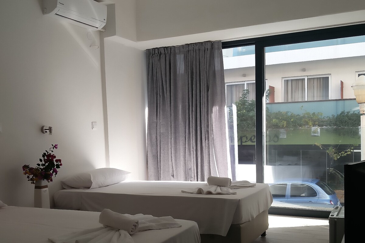 Habitación de hotel con dos camas, un ventanal con cortinas, aire acondicionado en el techo y vista a un edificio y un automóvil fuera.