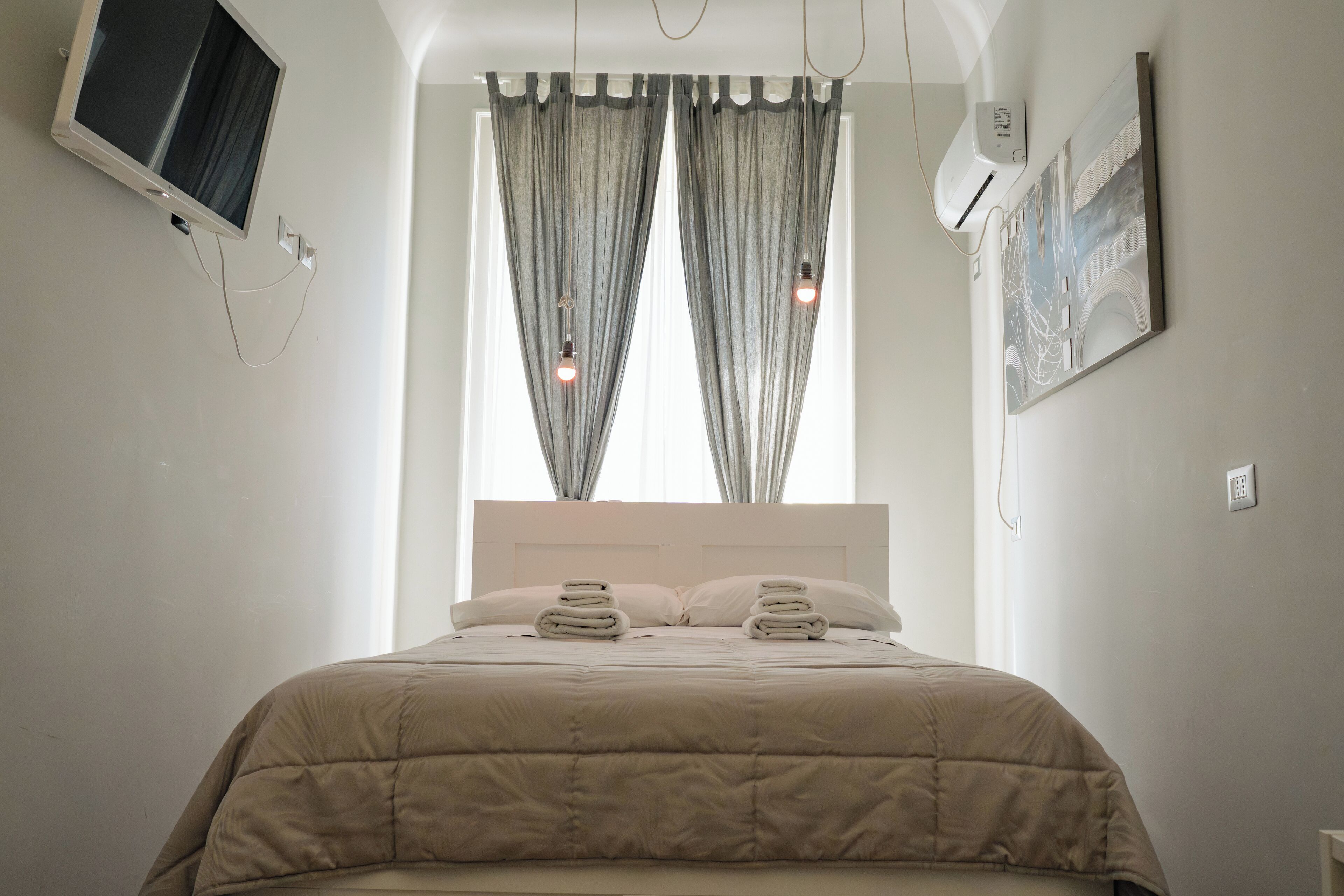 Een net opgemaakt bed met hoofdbord, een wandtelevisie en een schilderij aan de muur.