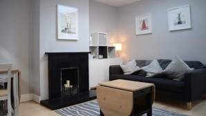 A modern living room with a fireplace, a black sofa, a wooden coffee table, and framed artwork on the walls. - Charming 2 Bedroom Cottage in Central Location (Dublin)