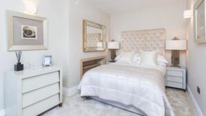 A bedroom with a large bed, a dresser, a nightstand, a mirror, and wall-mounted lamps. - Luxury Mayfair 2 Bedroom Apartment (London)