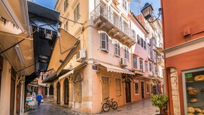 Una calle estrecha flanqueada por edificios con balcones, toldos y una tienda de productos de panadería.