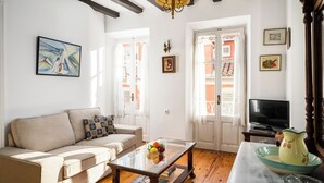 Una sala de estar con sofá, una mesa de centro y televisor. Hay ventanas con contraventanas, una araña y cuadros enmarcados en las paredes.
