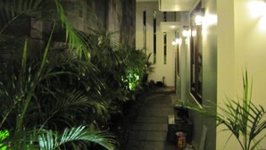 A narrow pathway flanked by lush green plants leading to a building with a white facade and multiple light fixtures. - Omahqu Guest House (Yogyakarta)
