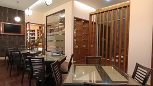 A dining area with a glass-top table and black chairs, a wooden cabinet with glass doors, and a TV mounted on the wall. - Omahqu Guest House (Yogyakarta)
