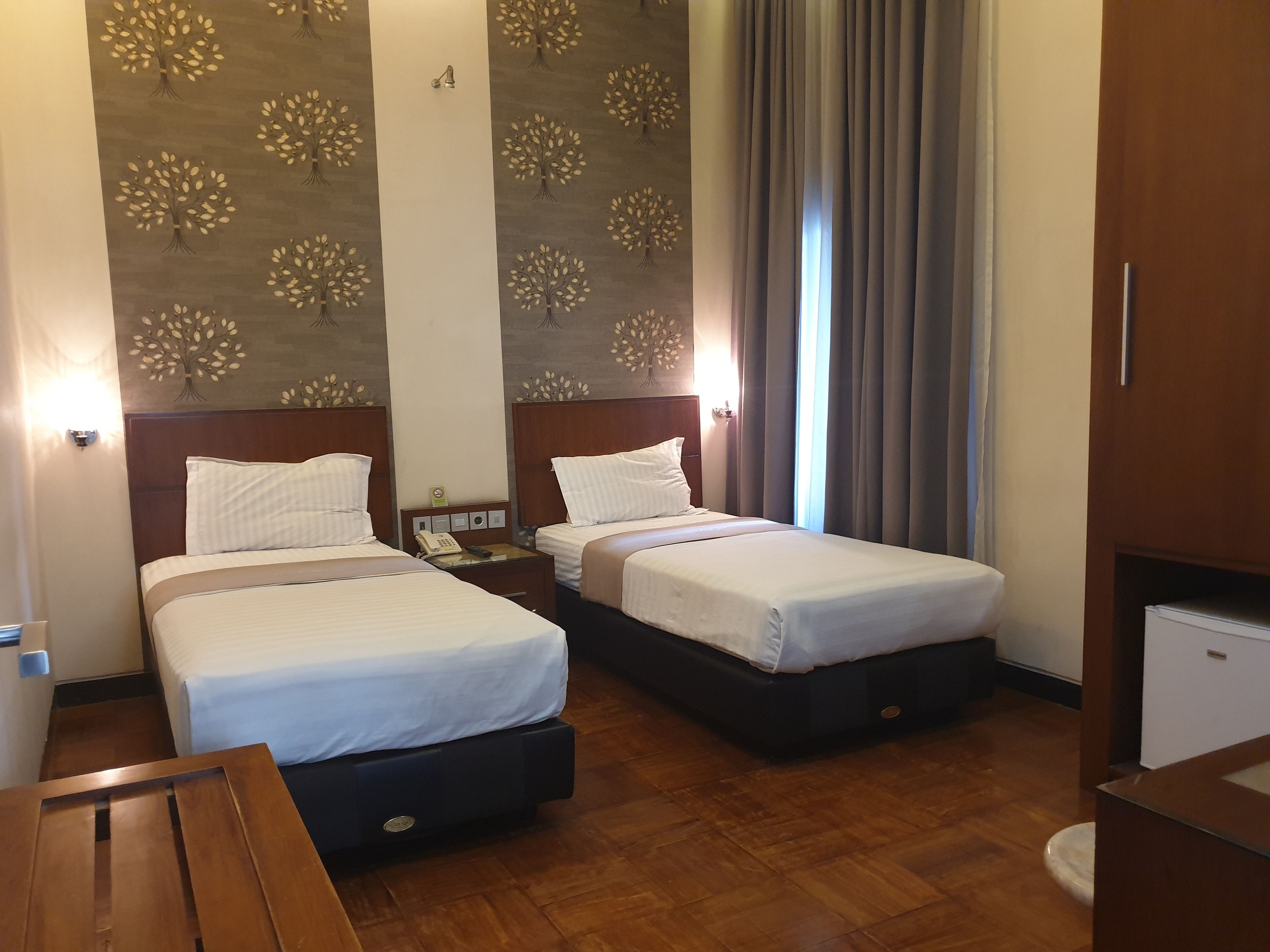 Quarto de hotel com duas camas, piso de madeira, uma secretária pequena e uma parede com padrão de árvore.