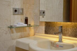 A bathroom with a marble wall, a glass shelf with toiletries, a sink with a faucet, and a mirror above the sink. - Omahqu Guest House (Yogyakarta)
