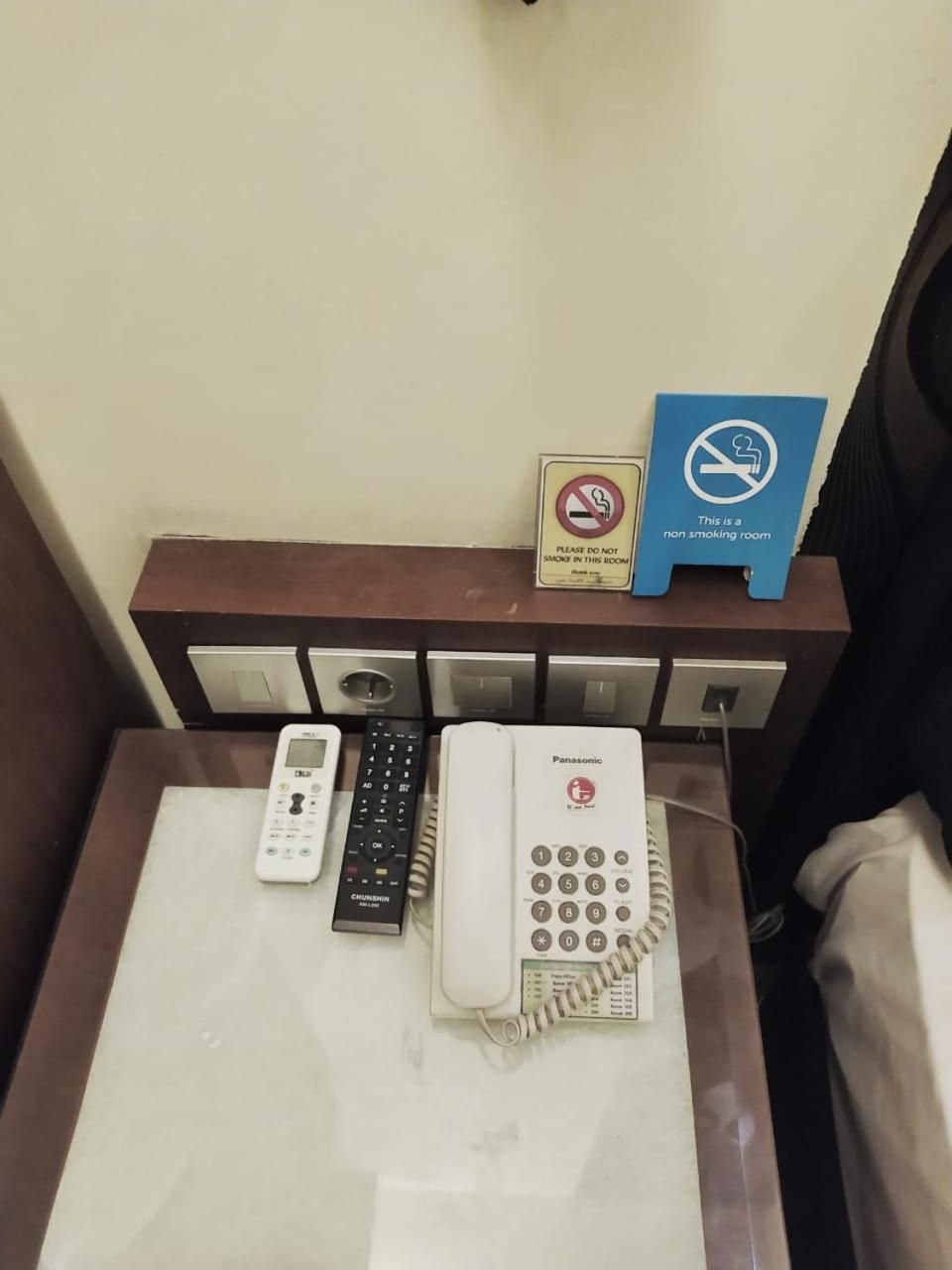 A hotel room with a bed, a bedside table with a phone, a remote control, and a non-smoking sign.
