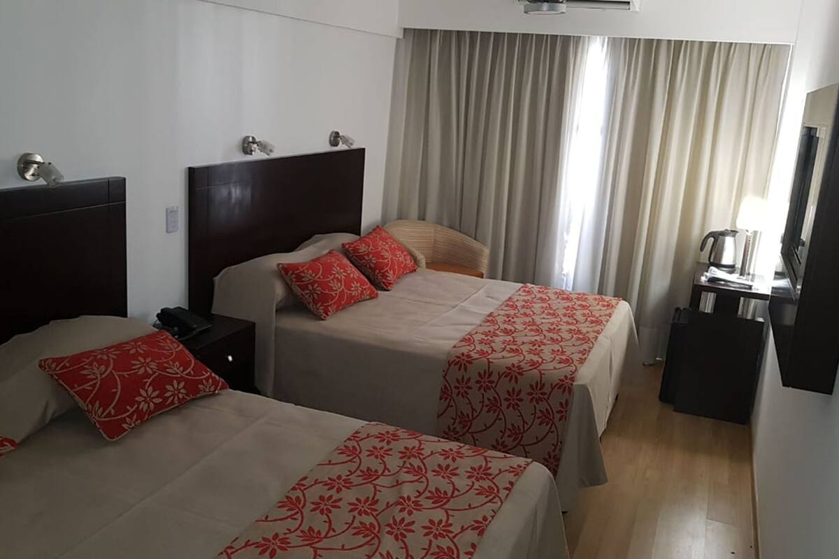 Habitación de hotel con dos camas, cada una con ropa de cama floral roja y cabeceros de madera. Hay un escritorio con cafetera y un espejo en la pared.