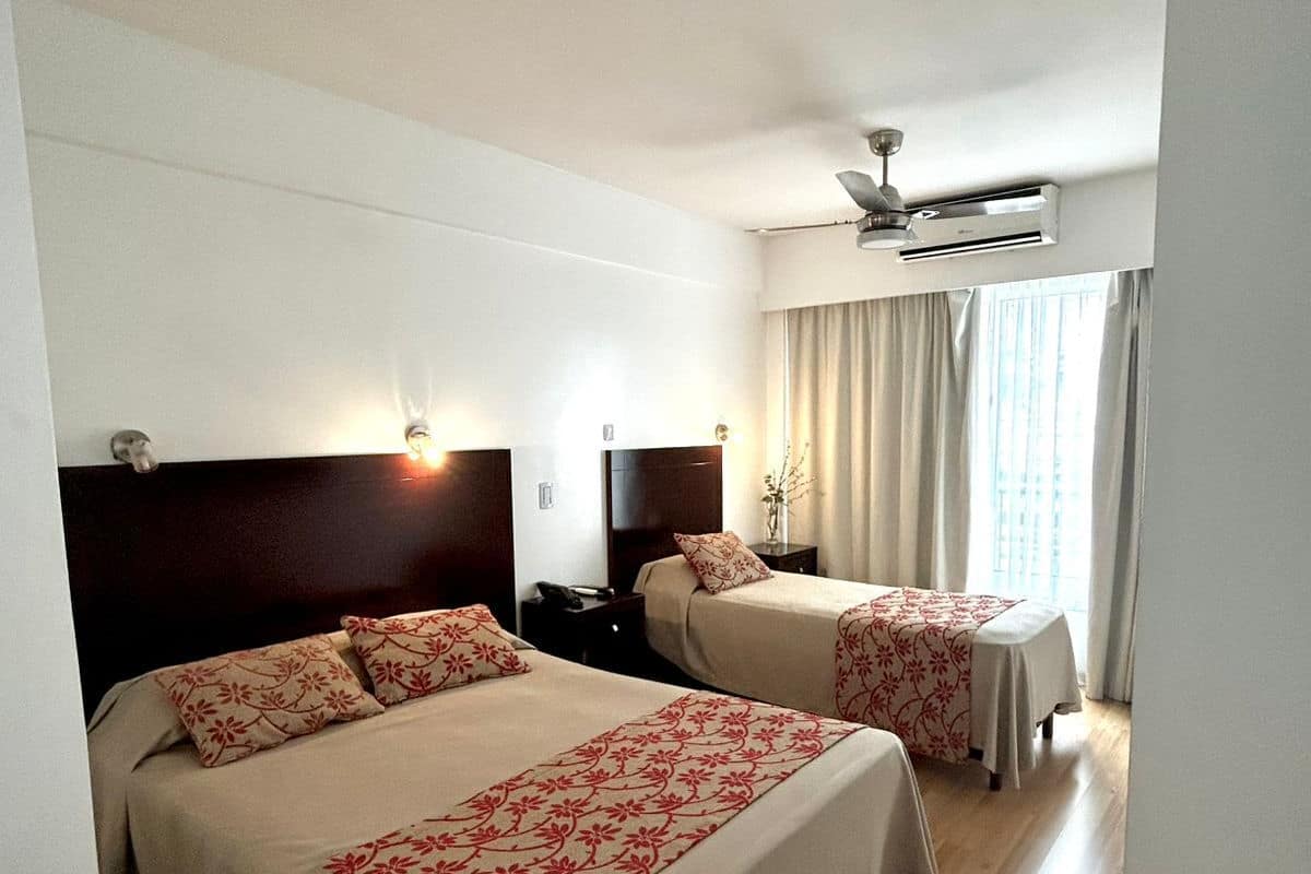 Habitación de hotel con dos camas, ventilador de techo y ventana con cortinas.