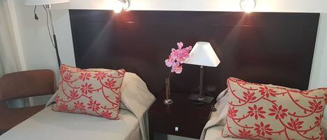 Une chambre d’hôtel avec deux lits, chacun agrémenté d’un oreiller à motifs floraux, une table de chevet avec une lampe et un vase contenant des fleurs roses.