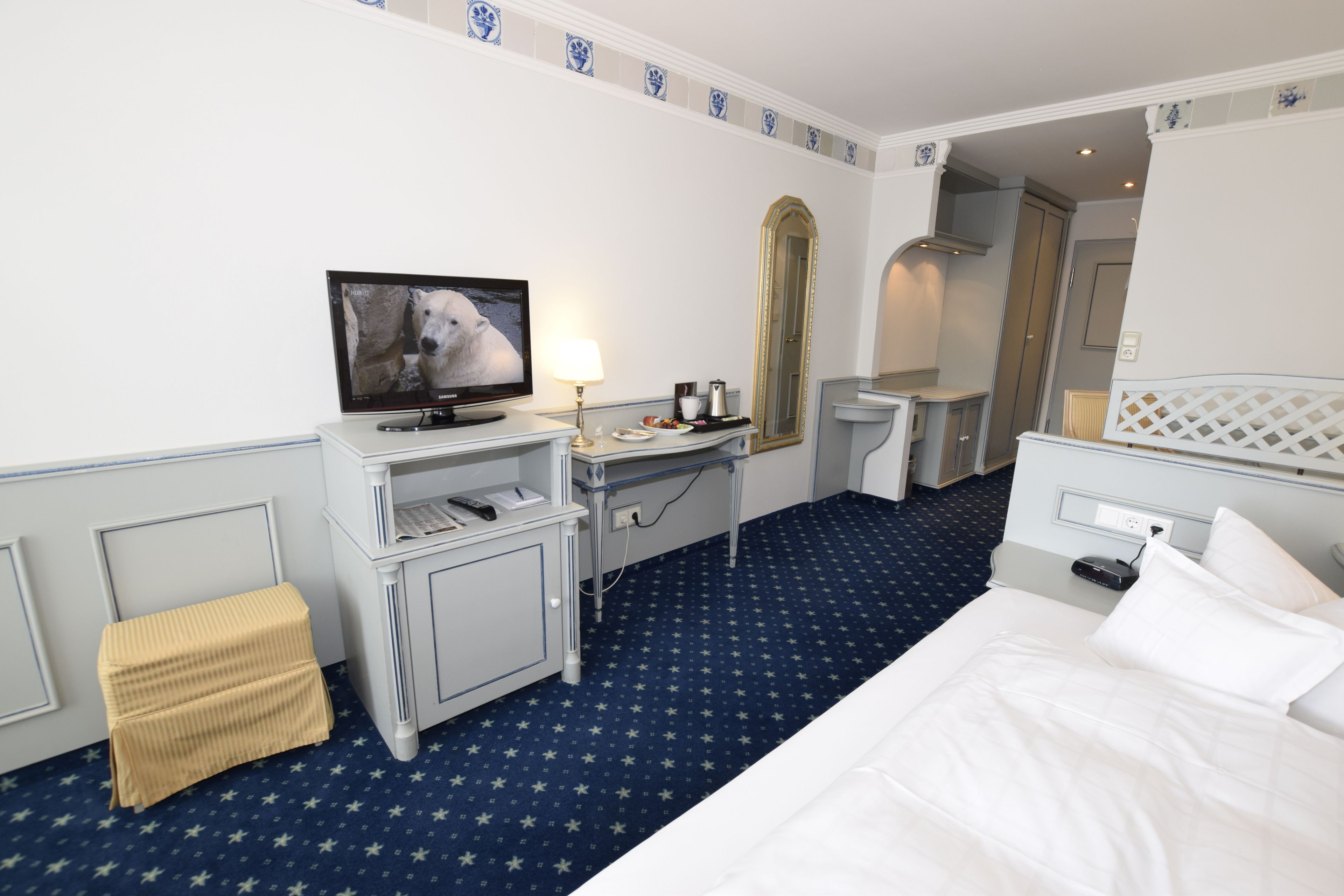 Habitación de hotel con cama, escritorio con televisor, espejo y un taburete pequeño.